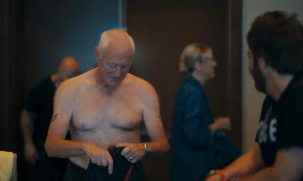 Barry Hearn shirtless, showing tattoos, with other people in the background.
