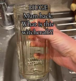 A hand holding a glass measuring cup filled with liquid over a soapy sink.