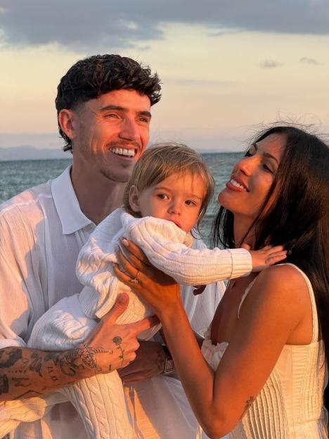 A man and woman smiling at each other while holding a baby against a sunset ocean backdrop.