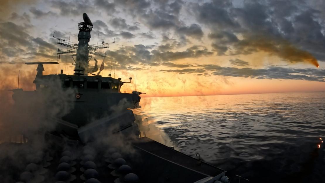 A Royal Navy warship fires a missile over the ocean at sunset.