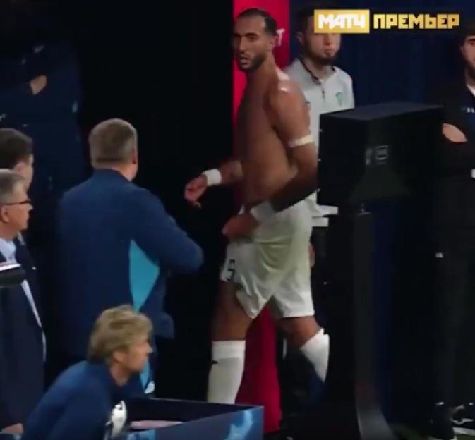 A shirtless soccer player in white shorts and white socks walking past people in a dark room.
