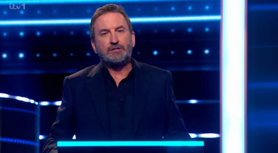 Screenshot of a man in a suit speaking on a game show set.