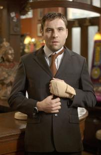 David Walliams as Ray in EastEnders, wearing a suit and a bandage on his hand.
