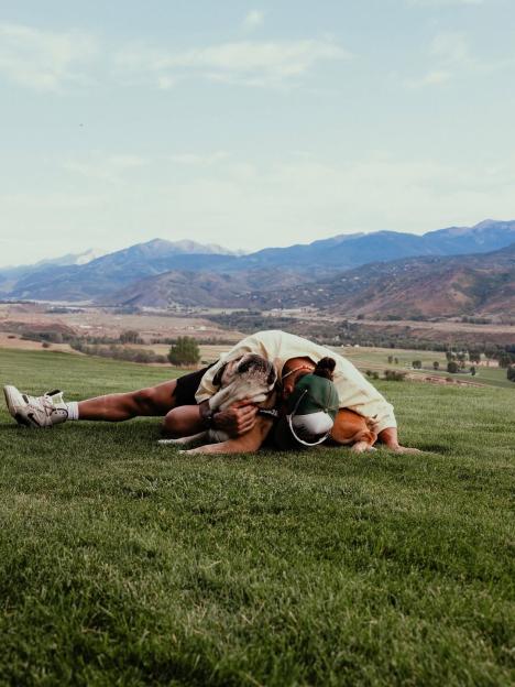 Person hugging a dog on a grassy field with mountains in the background.