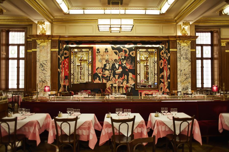 The interior of Brasserie Zédel, showing a large art deco mural, a red velvet banquette, and tables with pink and white tablecloths.