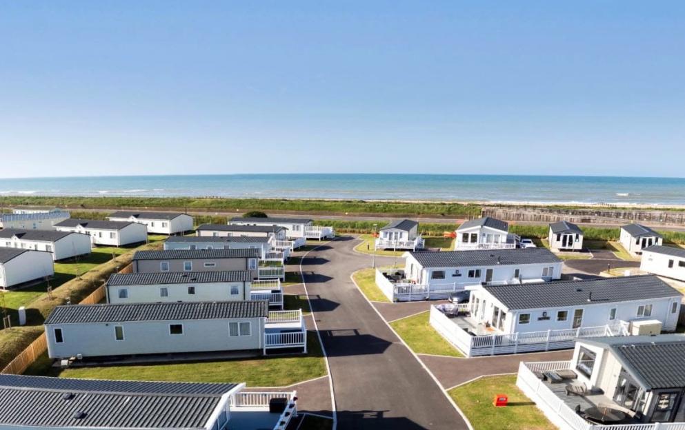 An aerial view of Camber Sands Holiday Park, showing rows of white holiday homes with dark grey roofs, green lawns, and paved roads leading towards the blue sea.