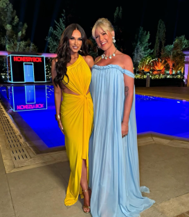 Two women in formal gowns posing by a pool with Honesty Box structures in the background.
