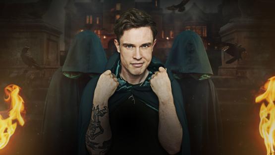 Ed Gamble in a green cloak, flanked by hooded figures, in front of a spooky mansion.