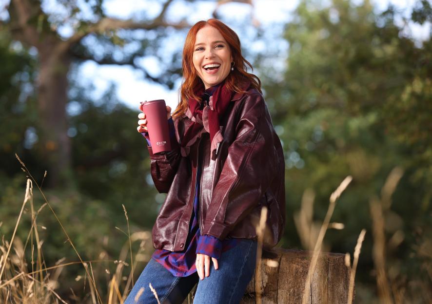 Angela Scanlon holding a Costa Coffee reusable cup while sitting on a tree stump in Epping Forest.