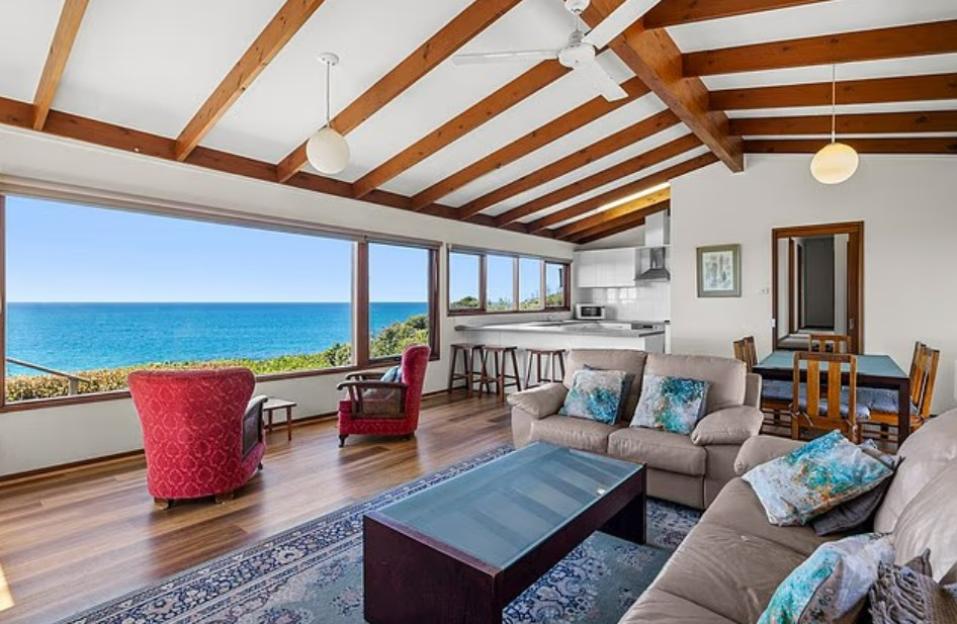 Interior of a house with an ocean view, showing a living room with two couches, a kitchen, and a dining area.