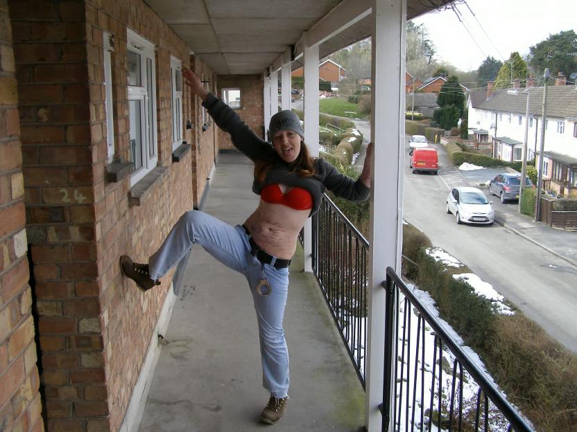 A woman posing playfully on a balcony, wearing a bra and jeans with handcuffs on her belt.
