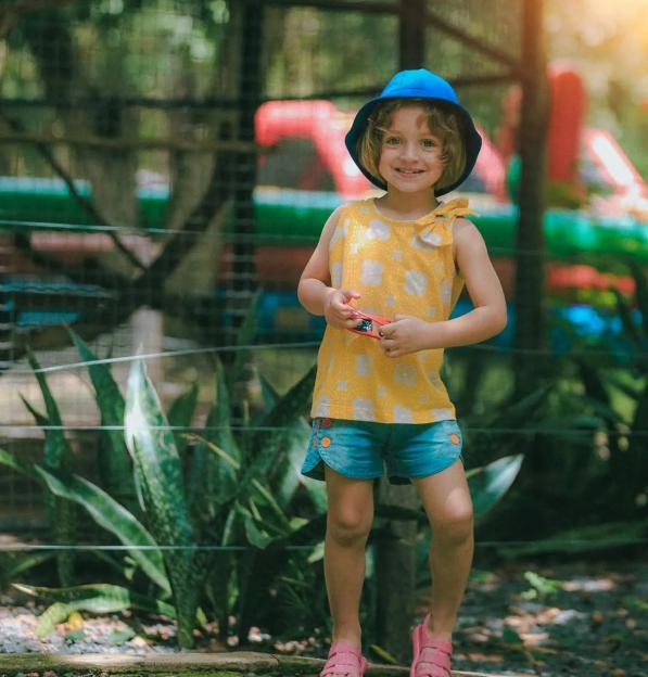 A smiling young girl in a blue hat and yellow shirt stands outdoors.