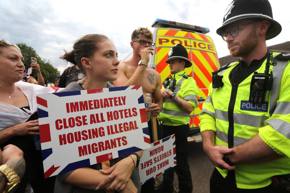 Protestors holding signs that read "IMMEDIATELY CLOSE ALL HOTELS HOUSING ILLEGAL MIGRANTS" at a demonstration, watched by police officers.