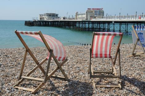 Wooden deck chairs on a pebble beach with a pier in the background.