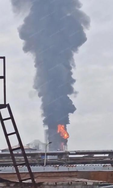 A fire with dense black smoke rising from an oil plant, seen over a partially visible structure with a metal ladder.