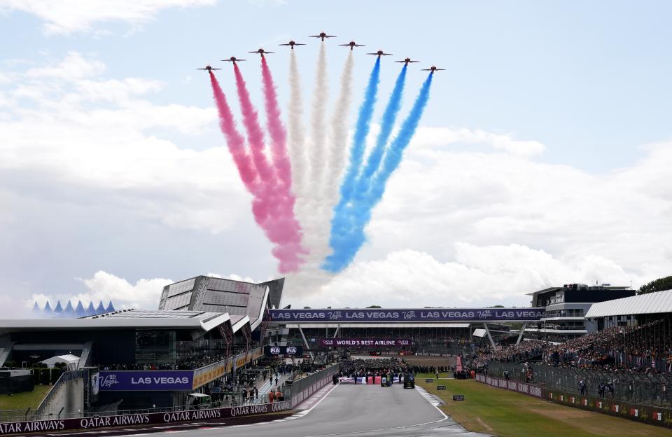 The Red Arrows performing a flypast, leaving red, white, and blue smoke trails over a racetrack filled with spectators.