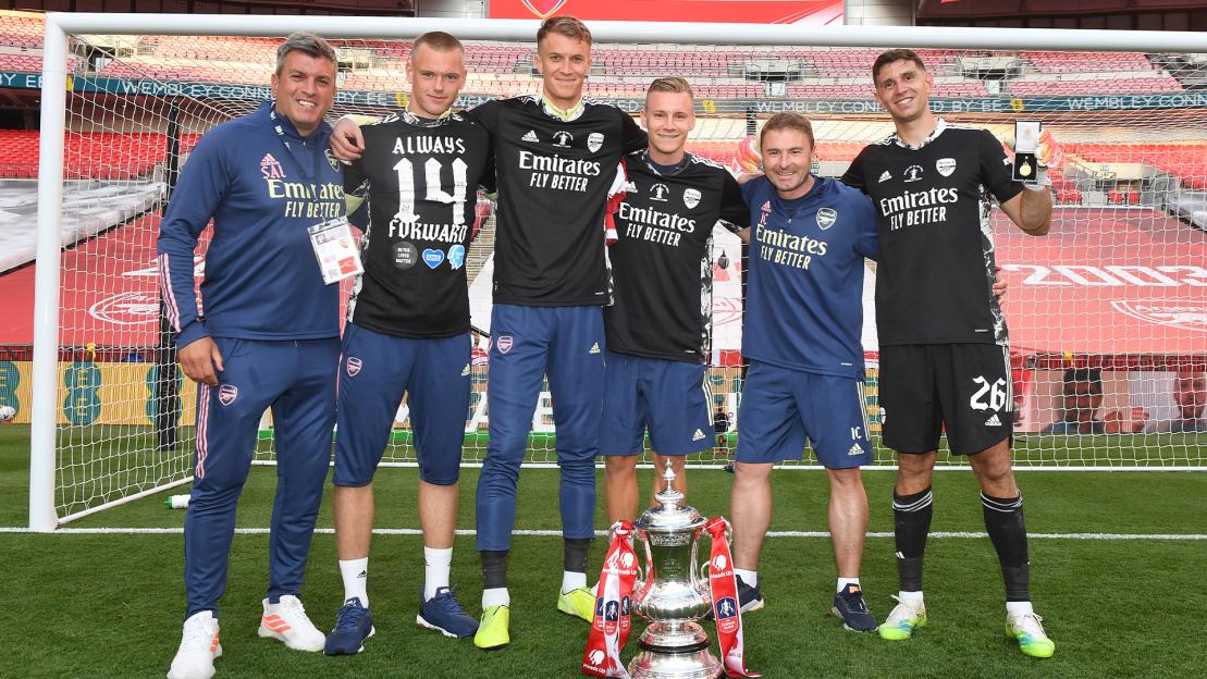 Six Arsenal FC players and staff pose with the FA Cup trophy on a soccer field.