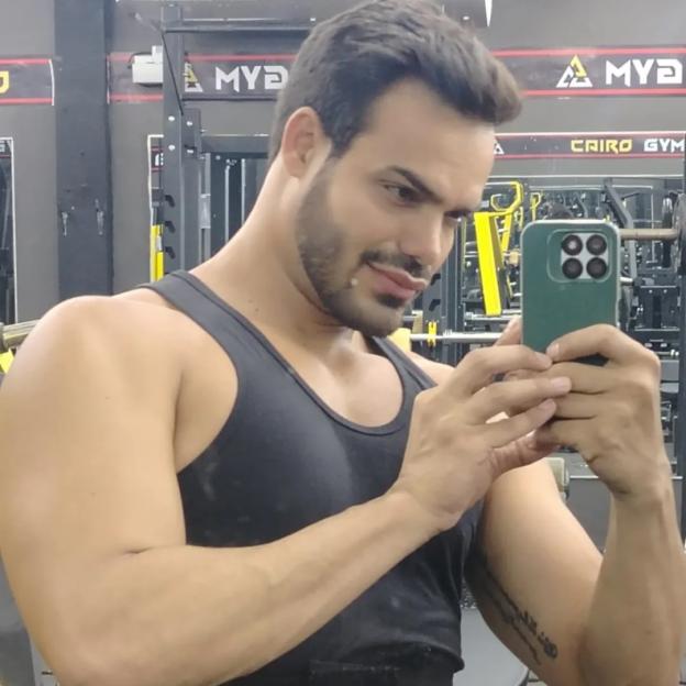 Emilio Sueños, a TV star and model, wearing a black tank top and taking a mirror selfie in a gym.