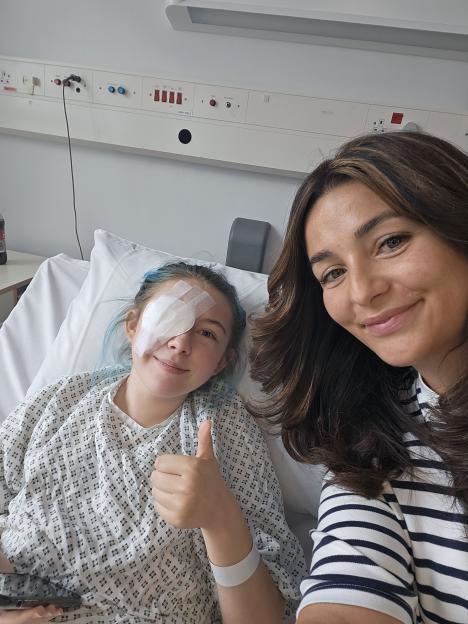Photo of a young woman in a hospital bed giving a thumbs-up, with her mother beside her.