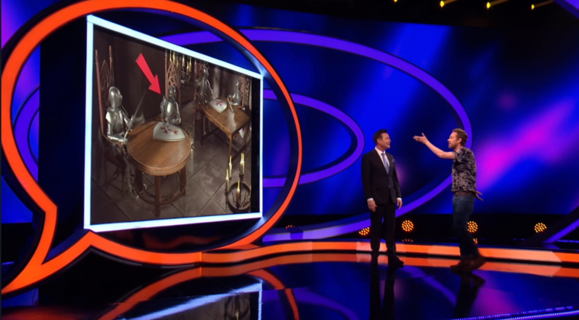 Screenshot of two men on a game show stage, one pointing at a screen showing two knights seated at a table.
