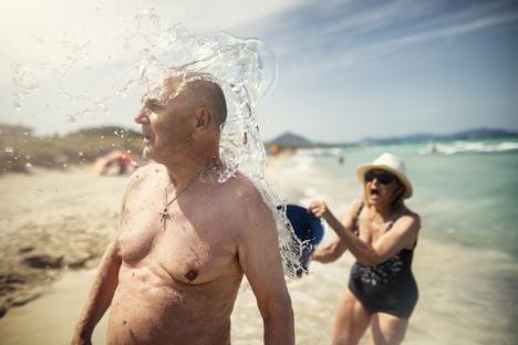 Senior woman surprising senior man with bucket of water on beach.