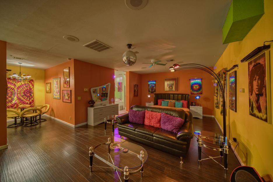Interior view of a brightly colored room with a leather couch, glass coffee table, and tie-dye decor.