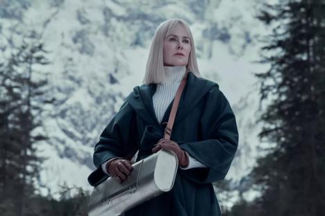 Woman in a green coat carrying a metal case in a snowy mountain landscape.