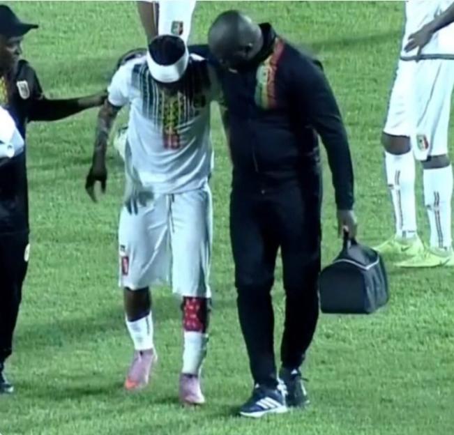 Injured soccer player wearing a white jersey being helped off the field by two staff members.