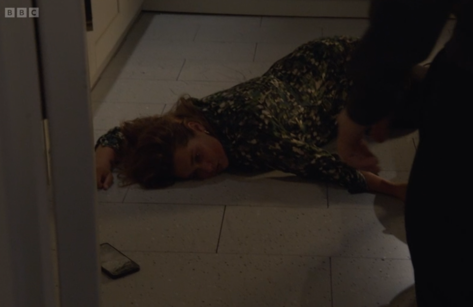 A woman in a printed dress is lying on a light-colored tiled floor, with her head resting on her left arm, and her eyes partially closed. A phone is on the floor near her left side, and a person's leg is visible on the right.