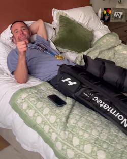 Man in bed wearing a marathon medal and recovery boots.