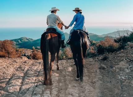 Two women holding hands while horseback riding overlooking the ocean.
