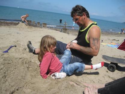 Photograph of a father and daughter on a beach; the father is adjusting the daughter's shoes.