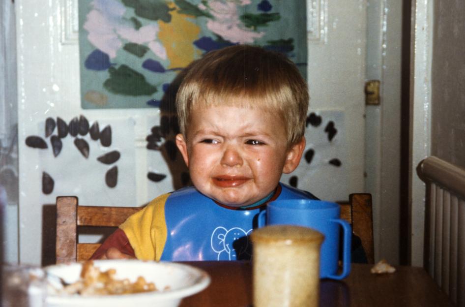 Andrew Gosden as a 2-year-old in a blue bib and yellow shirt, sitting at a table with a white bowl, crying.