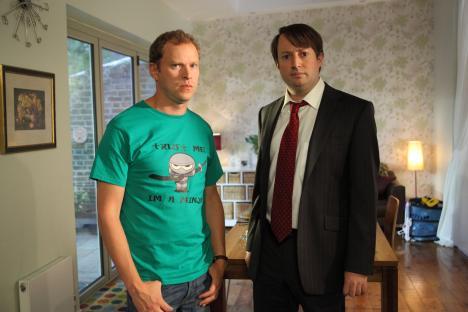 Two men standing in a living room; one wears a green t-shirt with a ninja illustration, the other a suit and tie.