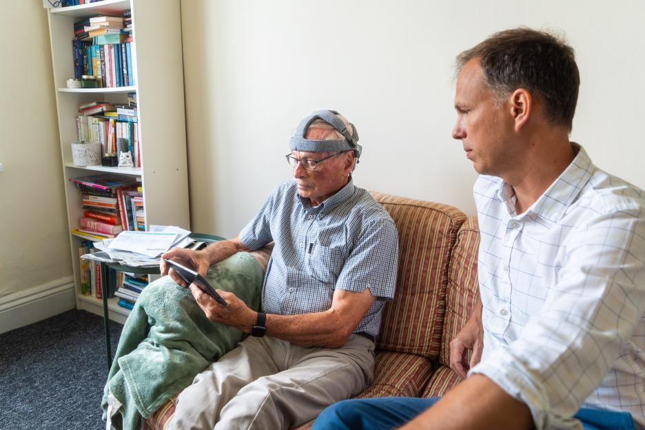 Older man wearing a brainwave-reading device, sitting on a couch with a younger man.