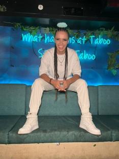 Woman with long braids sitting on a teal couch in front of a blue wall with neon lights.