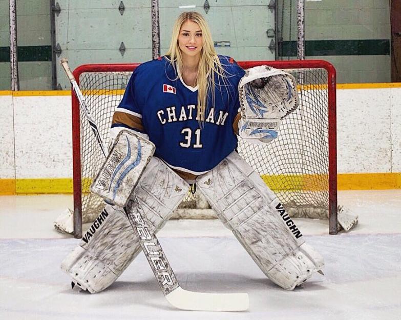 Mikayla Demaiter, a female hockey goalie, wearing her goalie gear and posing on the ice in front of a hockey net.