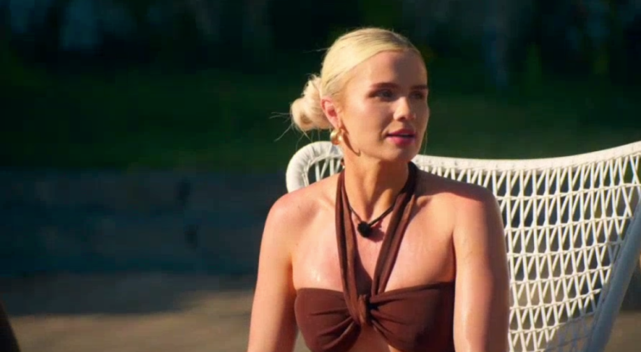 A woman with blonde hair in a bun, wearing a brown halter top, sitting in a white chair.