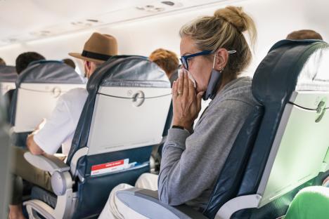 Woman blowing her nose on a plane with her face mask under her chin.