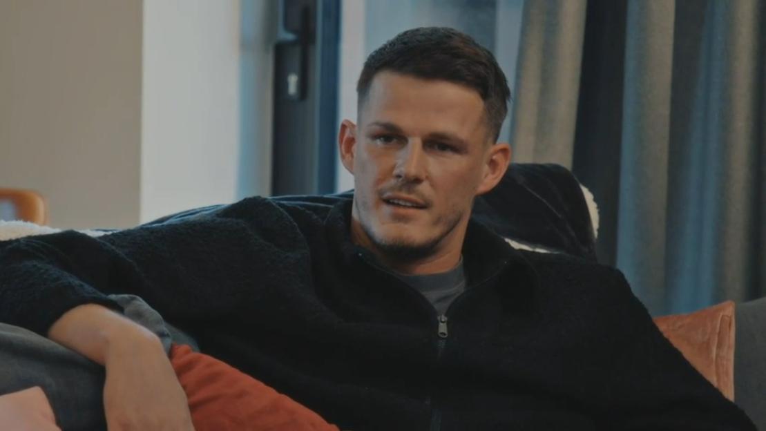 A man in a black zipped jacket sits on a couch, appearing concerned.