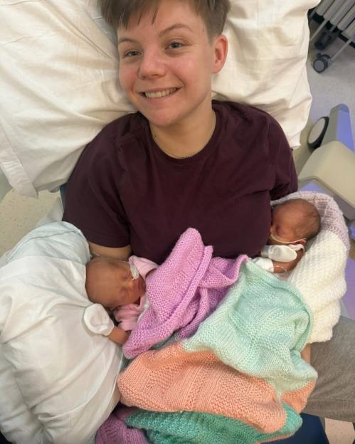 A smiling person holding twin newborns wrapped in pastel blankets.