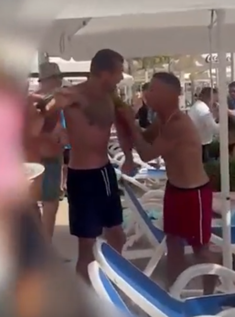 Two men in swim shorts fighting by a poolside.