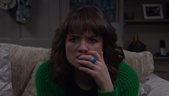 Screenshot of a woman with brown hair wearing a green sweater, looking anxious.
