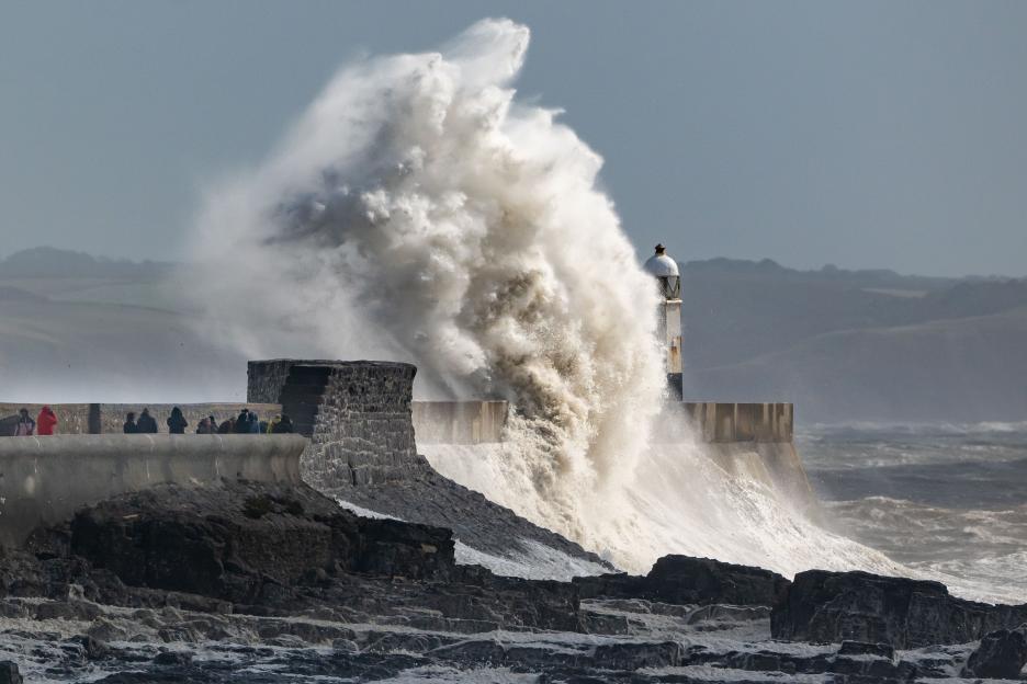 Large waves crashing against a harbor wall with a lighthouse in the background.