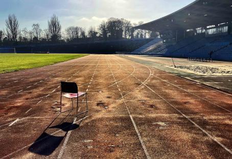 A single chair sits on a weathered running track in front of an empty stadium.