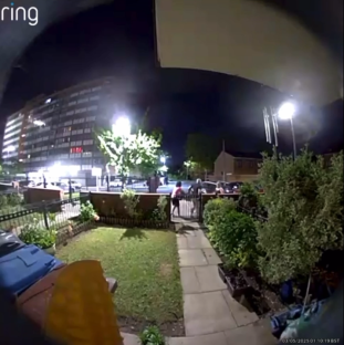 Nighttime security camera footage of a person opening a gate and walking toward a house.