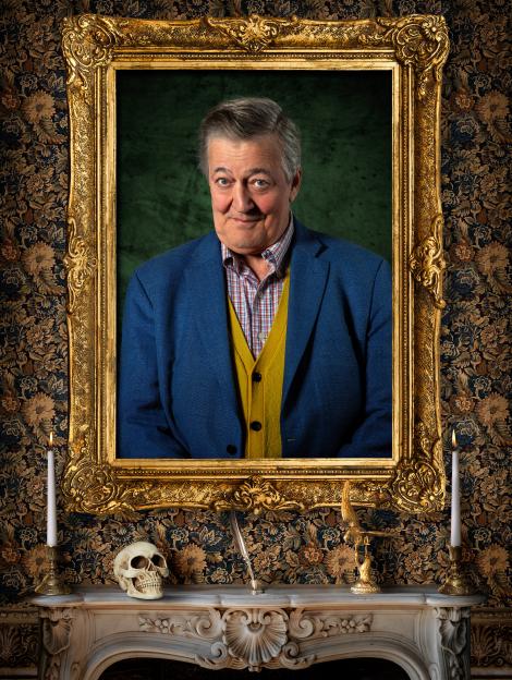 Stephen Fry in a gold ornate frame above a mantelpiece with a skull, quill, and two candles.