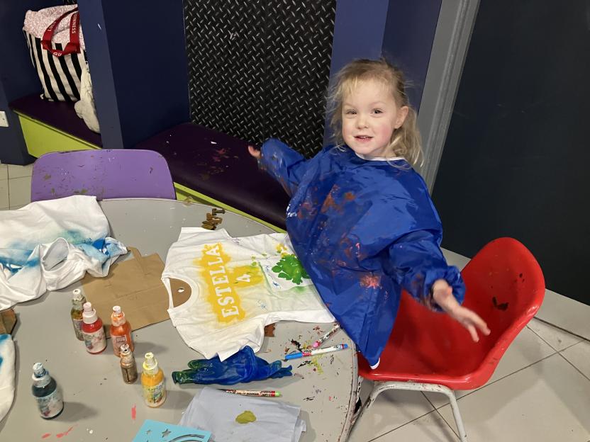A young girl in a blue paint smock sits at a table, decorating a t-shirt.