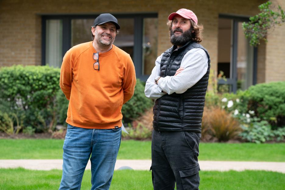 Joe Wilkinson in an orange sweatshirt and David Earl in a black vest in a garden.