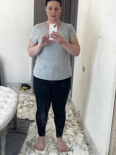Woman in gray shirt and black pants taking a selfie in a bathroom mirror.